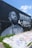 A large mural painted on a black wall features a monochrome portrait of a man with braided hair and a beard. The words 'Hustle & Motivate' appear in stylized lettering next to the portrait. The ground in front of the wall is covered with various colorful graffiti and signatures. Sparse patches of green grass can be seen along the edges.