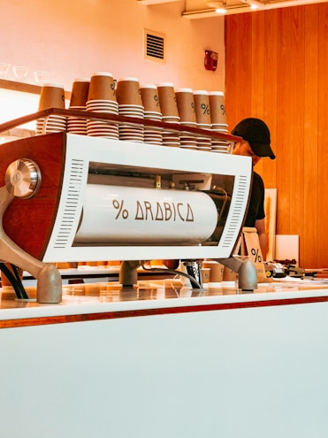 A stylish coffee counter featuring a modern espresso machine branded with 'Arabica'. Several stacks of brown paper cups are arranged on top of the machine. Wooden paneling and soft lighting create a cozy and inviting atmosphere, with a partial view of a person behind the counter.