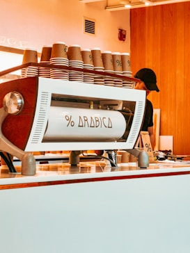 A stylish coffee counter featuring a modern espresso machine branded with 'Arabica'. Several stacks of brown paper cups are arranged on top of the machine. Wooden paneling and soft lighting create a cozy and inviting atmosphere, with a partial view of a person behind the counter.