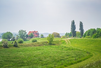 A picturesque rural landscape features a red-brick house with a sloped roof nestled among lush trees and greenery. A winding dirt path curves through a vibrant green field, leading towards the house. Tall, slender trees punctuate the horizon, and a few people can be seen walking along the path in the background. The scene is serene and bathed in soft, natural light.