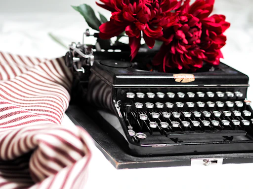 A moody black-and-white photo of a vintage typewriter with a single red rose beside it, evoking a noir poetic atmosphere.