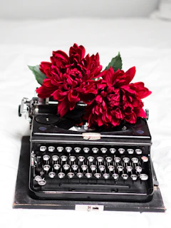A serene black and white photograph of a vintage typewriter on a wooden desk, with a single red carnation beside it.