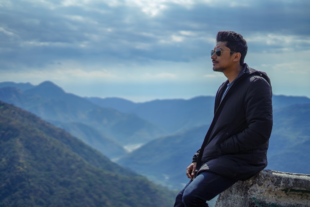 검은 재킷을 입은 남자가 낮에 산을 바라보며 바위에 앉아 있다