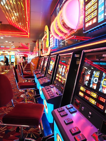 A brightly lit casino interior featuring a row of colorful slot machines. The neon lights create a vibrant atmosphere, with patterned carpeting adding to the decor. Stools are aligned neatly in front of each machine.