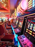 A brightly lit casino interior featuring a row of colorful slot machines. The neon lights create a vibrant atmosphere, with patterned carpeting adding to the decor. Stools are aligned neatly in front of each machine.