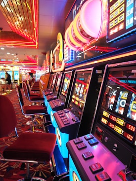 A brightly lit casino interior featuring a row of colorful slot machines. The neon lights create a vibrant atmosphere, with patterned carpeting adding to the decor. Stools are aligned neatly in front of each machine.