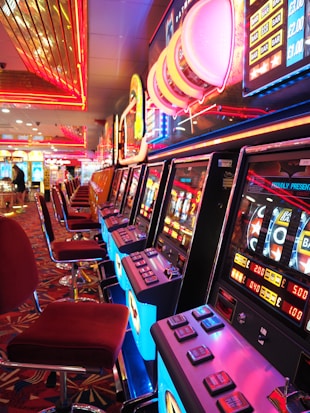 A brightly lit casino interior featuring a row of colorful slot machines. The neon lights create a vibrant atmosphere, with patterned carpeting adding to the decor. Stools are aligned neatly in front of each machine.