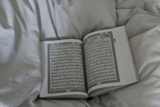 An open book displaying Arabic script, possibly a religious text, rests on a soft, white bedspread. The surrounding fabric appears plush and inviting.