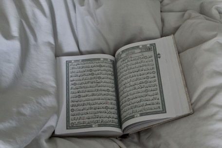 An open book displaying Arabic script, possibly a religious text, rests on a soft, white bedspread. The surrounding fabric appears plush and inviting.