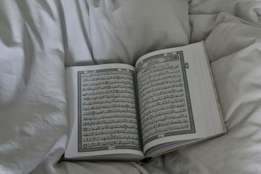 An open book displaying Arabic script, possibly a religious text, rests on a soft, white bedspread. The surrounding fabric appears plush and inviting.