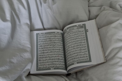 An open book displaying Arabic script, possibly a religious text, rests on a soft, white bedspread. The surrounding fabric appears plush and inviting.