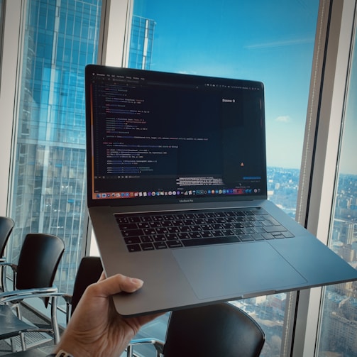 A person holds a laptop displaying code, with a high-rise cityscape visible through large windows in the background. The room contains several empty chairs, indicating a possible conference or meeting setting.
