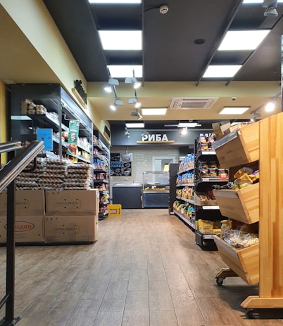 A well-lit grocery store aisle with shelves stocked with various goods, including packaged food items and eggs. Wooden bins show more items on the right side, and signs with Cyrillic text are visible towards the back.