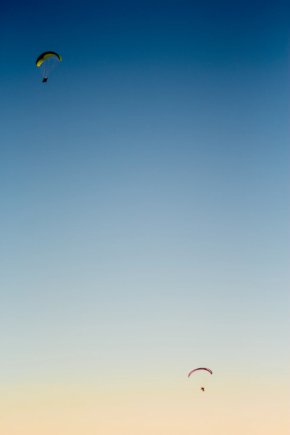 Two people paraglide in colorful parachutes against a clear blue sky with wispy clouds