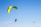 Two paragliders are flying in the sky, both featuring bright green parachutes against a clear blue background. One paraglider is larger and closer, with visible harness and equipment. The other is smaller and appears further away.