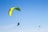 Two paragliders are flying in the sky, both featuring bright green parachutes against a clear blue background. One paraglider is larger and closer, with visible harness and equipment. The other is smaller and appears further away.