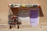 A rustic building with a light purple door set into a sandy brown wall. There's a colorful fabric awning above the door, and a metal rack to the left holding both red and black spherical objects. A sign in Arabic is painted on the wall above the rack and to the right of the door.