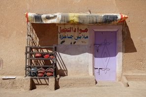 A rustic building with a light purple door set into a sandy brown wall. There's a colorful fabric awning above the door, and a metal rack to the left holding both red and black spherical objects. A sign in Arabic is painted on the wall above the rack and to the right of the door.