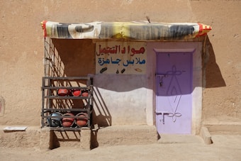 A rustic building with a light purple door set into a sandy brown wall. There's a colorful fabric awning above the door, and a metal rack to the left holding both red and black spherical objects. A sign in Arabic is painted on the wall above the rack and to the right of the door.