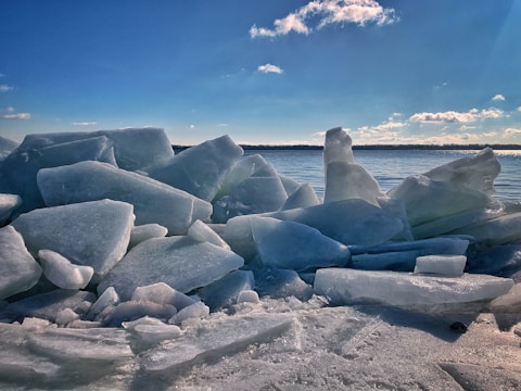 Large chunks of ice are piled along the shore of a body of water under a clear blue sky with some clouds. The ice pieces are irregularly shaped and appear to be a mixture of transparent and opaque surfaces. In the distance, there is a visible treeline across the water.