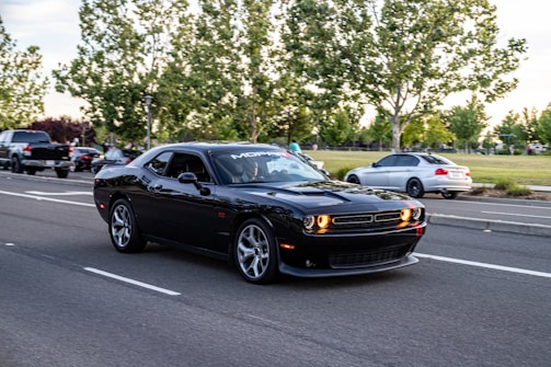 A black modern muscle car is prominently displayed on a suburban road, surrounded by several other vehicles. The car features a sleek design and bold, lit-up headlights. Tall green trees line the street, and a grassy park area is visible in the background. The scene is captured during daylight with a clear sky.