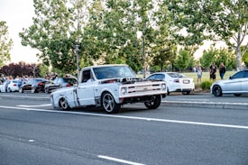 A white and heavily modified old pickup truck is driving on a road, with several parked cars on the side. There are people walking and standing near the parked cars, and large green trees in the background, with some grass and shrubs. The pickup truck has visible engine modifications and large black rims.