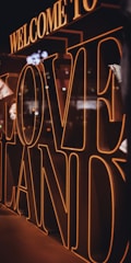 Large 3D letters spell out 'Welcome to Love Land' with warm lighting creating a glowing effect. The letters dominate the scene with reflections visible on their surfaces, suggesting an indoor environment.