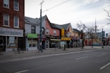 A street view featuring a row of storefronts and businesses with various signs in English and Chinese. The architecture includes brick buildings and some older, traditional designs. There is minimal activity with no visible people, and a few bicycles are parked along the sidewalk. Leafless trees line the street, and the CN Tower is visible in the background.
