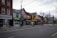 A street view featuring a row of storefronts and businesses with various signs in English and Chinese. The architecture includes brick buildings and some older, traditional designs. There is minimal activity with no visible people, and a few bicycles are parked along the sidewalk. Leafless trees line the street, and the CN Tower is visible in the background.