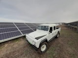 A rugged white pickup truck is parked on a patch of grass next to a large array of solar panels, which stretch into the distance under a cloudy sky.