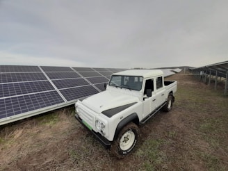 A rugged white pickup truck is parked on a patch of grass next to a large array of solar panels, which stretch into the distance under a cloudy sky.