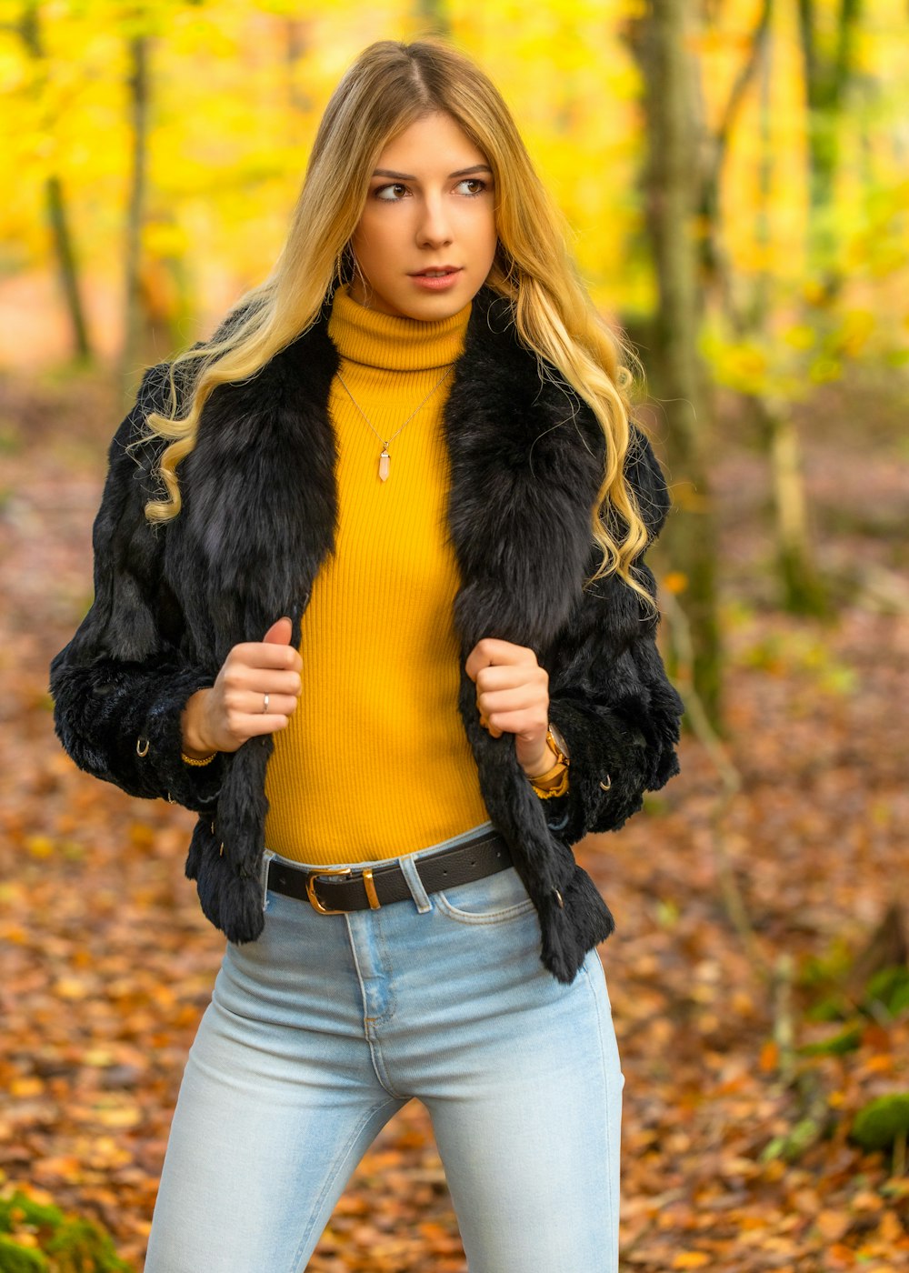 woman in black fur jacket and blue denim jeans standing on dried leaves during daytime