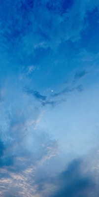 A serene sky with varying shades of blue, featuring scattered clouds and a small crescent moon at the center. The subtle gradient from deeper blue at the top to lighter blue at the horizon creates a calming effect.