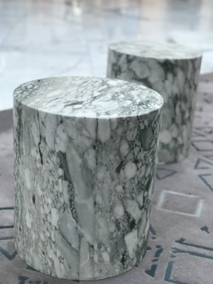Two cylindrical stools made of marble with a blend of white and grey tones are placed on a textured floor with geometric patterns.