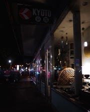 A dimly lit street scene at night features the storefront of a shop with a sign reading 'Kyoto'. The display inside the window shows various items, possibly decorative or cultural artifacts, subtly illuminated. The sidewalk outside is dark, with some streetlights and illuminated signs further along the path, creating a sense of quiet urban atmosphere.