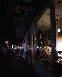 A dimly lit street scene at night features the storefront of a shop with a sign reading 'Kyoto'. The display inside the window shows various items, possibly decorative or cultural artifacts, subtly illuminated. The sidewalk outside is dark, with some streetlights and illuminated signs further along the path, creating a sense of quiet urban atmosphere.