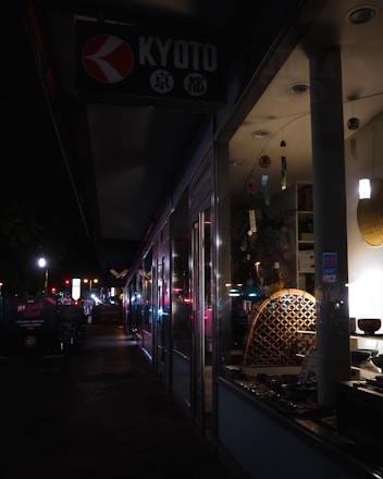 A dimly lit street scene at night features the storefront of a shop with a sign reading 'Kyoto'. The display inside the window shows various items, possibly decorative or cultural artifacts, subtly illuminated. The sidewalk outside is dark, with some streetlights and illuminated signs further along the path, creating a sense of quiet urban atmosphere.