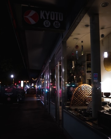 A dimly lit street scene at night features the storefront of a shop with a sign reading 'Kyoto'. The display inside the window shows various items, possibly decorative or cultural artifacts, subtly illuminated. The sidewalk outside is dark, with some streetlights and illuminated signs further along the path, creating a sense of quiet urban atmosphere.