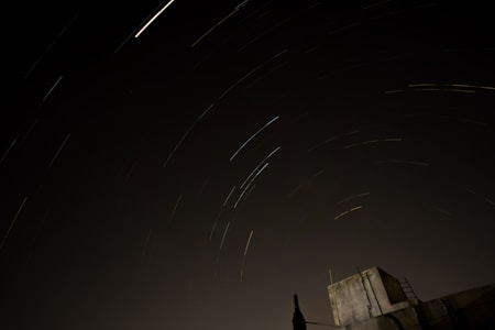 A long exposure photograph captures the trails of stars in the night sky, creating smooth, curved streaks of light across a dark backdrop. In the foreground is the silhouette of a structure, possibly a rooftop, with a ladder and antenna visible against the sky.