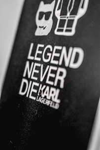 A black background features bold white text stating 'Legend Never Die' followed by the name 'Karl Lagerfeld.' Above the text, there are simple white line drawings of a cat wearing sunglasses and a stylized human figure.
