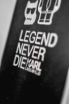 A black background features bold white text stating 'Legend Never Die' followed by the name 'Karl Lagerfeld.' Above the text, there are simple white line drawings of a cat wearing sunglasses and a stylized human figure.