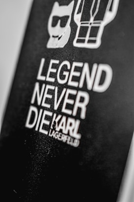 A black background features bold white text stating 'Legend Never Die' followed by the name 'Karl Lagerfeld.' Above the text, there are simple white line drawings of a cat wearing sunglasses and a stylized human figure.