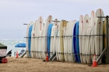 A row of surfboards is neatly lined up on a rack on a sandy beach. Some surfboards are decorated with logos and have colorful designs. In the background, there are a few beach umbrellas, and the ocean is visible with gentle waves.