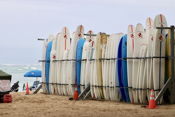 A row of surfboards is neatly lined up on a rack on a sandy beach. Some surfboards are decorated with logos and have colorful designs. In the background, there are a few beach umbrellas, and the ocean is visible with gentle waves.