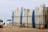 A row of surfboards is neatly lined up on a rack on a sandy beach. Some surfboards are decorated with logos and have colorful designs. In the background, there are a few beach umbrellas, and the ocean is visible with gentle waves.
