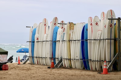 A row of surfboards is neatly lined up on a rack on a sandy beach. Some surfboards are decorated with logos and have colorful designs. In the background, there are a few beach umbrellas, and the ocean is visible with gentle waves.
