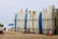 A row of surfboards is neatly lined up on a rack on a sandy beach. Some surfboards are decorated with logos and have colorful designs. In the background, there are a few beach umbrellas, and the ocean is visible with gentle waves.