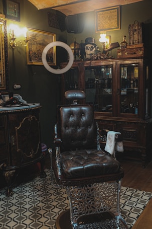 A vintage-style barber shop interior with a dark leather barber chair positioned centrally. The room is decorated with ornate wooden cabinets and framed pictures on dark green walls. A skull adorned with a top hat and goggles is displayed on a cabinet, accompanied by various grooming products and retro lighting fixtures.