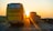 A yellow bus and a truck are traveling down a highway during sunset. The sun is low on the horizon, casting warm golden light across the scene. The road is bordered by greenery and there is a clear sky above.
