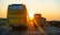 A yellow bus and a truck are traveling down a highway during sunset. The sun is low on the horizon, casting warm golden light across the scene. The road is bordered by greenery and there is a clear sky above.